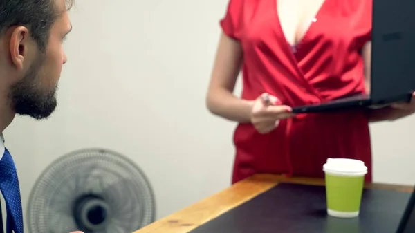 Office flirt. Een aantrekkelijke vrouw in een rode jumpsuit zet haar laptop op het Bureau van haar medemens. — Stockfoto