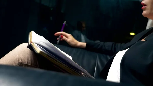 Primer plano. una mujer habla con alguien y toma notas en un cuaderno mientras está sentada por la noche en una silla de cuero negro en la oficina. El concepto de consulta, negociación . —  Fotos de Stock