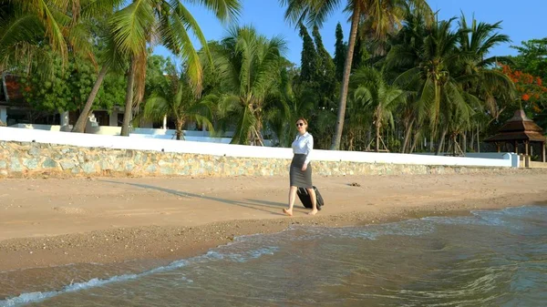 Επιχείρηση γυναίκα σε ρούχα γραφείου με μια βαλίτσα περπατάει ξυπόλητος κατά μήκος μιας λευκής αμμουδερές παραλία με φόντο φοίνικες και ένα πολυτελές θέρετρο. ανεξάρτητος, πολυαναμενόμενος σχεδιασμός αναψυχής — Φωτογραφία Αρχείου