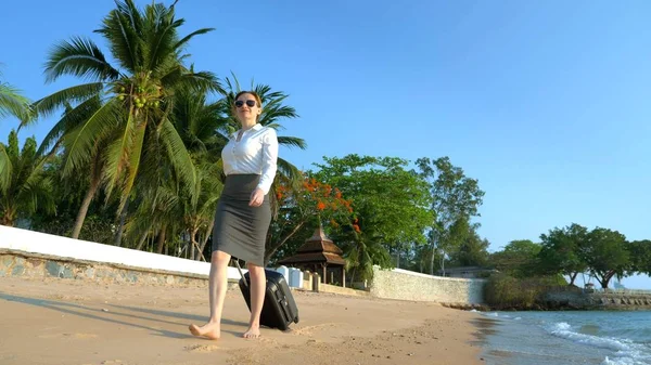Geschäftsfrau in Bürokleidung mit Koffer läuft barfuß an einem weißen Sandstrand vor der Kulisse von Palmen und einem Luxus-Resort entlang. freiberufliches, langersehntes Freizeitkonzept — Stockfoto