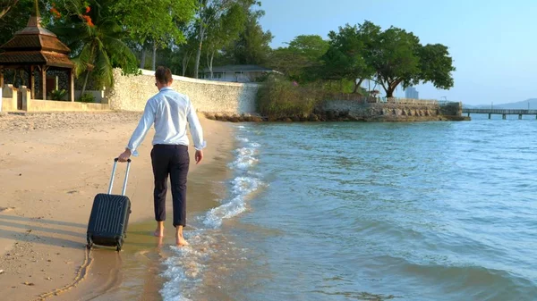 Hombre de negocios guapo en gafas de sol con una maleta va descalzo en la playa de arena blanca contra el telón de fondo de palmeras y un complejo de lujo. freelance, concepto de ocio largamente esperado — Foto de Stock