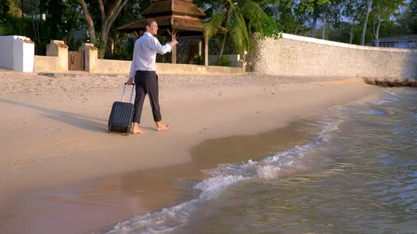 Bonito empresário de óculos de sol com uma mala vai descalço na praia de areia branca contra o pano de fundo de palmeiras e um resort de luxo. freelance, conceito de lazer tão esperado — Fotografia de Stock