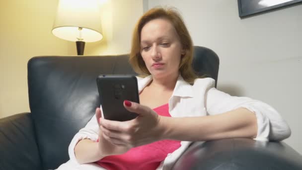 Egy nő használ egy okostelefon otthon, ül egy nagy bőr szék este, a háttérben egy állólámpa — Stock videók