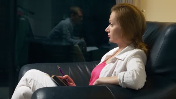 Close-up. een vrouw praat met iemand en neemt notities in een notitieblok zittend in de avond in een zwart lederen stoel in het kantoor. Het concept van overleg, onderhandelen. — Stockvideo
