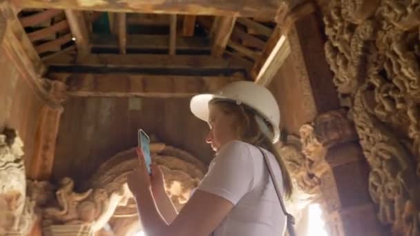 Vrouw toerist in een helm maakt een foto op haar smartphone in een houten boeddhistische tempel. — Stockvideo