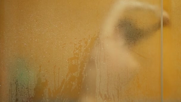 Naakt meisje wast in de douche. Haar vervaagde silhouet is zichtbaar door het oppervlak van het zweterige glas met druppels water. Vervagen — Stockvideo