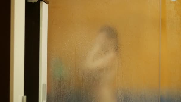 Naakt meisje wast in de douche. Haar vervaagde silhouet is zichtbaar door het oppervlak van het zweterige glas met druppels water. Vervagen — Stockvideo