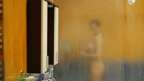 裸体女孩在淋浴时洗。她模糊的轮廓透过满是水滴的满是汗水的玻璃表面可见。模糊 — 图库视频影像
