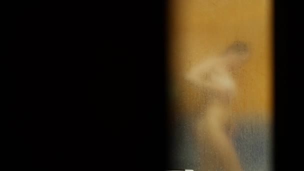 Jemand guckt durch die geöffnete Tür, während sich ein nacktes Mädchen unter der Dusche wäscht. Ihre verschwommene Silhouette ist durch die Oberfläche des verschwitzten Glases mit Wassertropfen sichtbar. Unschärfe — Stockvideo