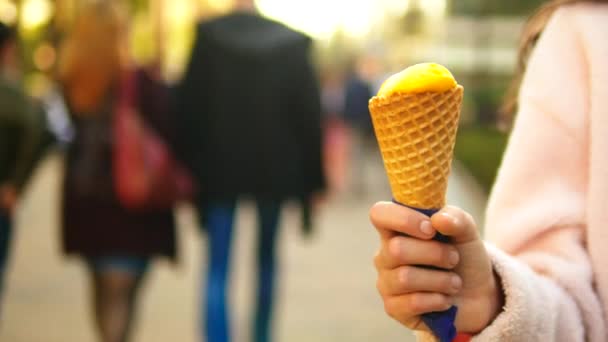 誰かがウエハーコーンにフルーツアイスクリームを保持し、クローズアップ。春または夏にぼやけた歩行者通りに対して — ストック動画