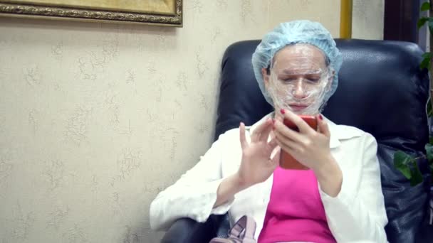 Uma mulher está lendo algo em um telefone celular, sentada em uma cadeira no corredor de uma clínica de cosmetologia com um filme para anestesia em seu rosto . — Vídeo de Stock