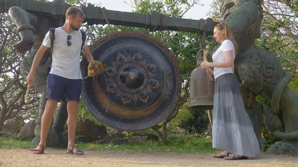 Sepasang turis seorang pria dan seorang wanita memakai helm sedang mengetuk sebuah gong tradisional Buddha pada perjalanan di sebuah kuil Buddha — Stok Foto