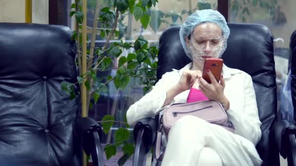 Kobieta czyta coś w telefonie komórkowym, siedząc na krześle w korytarzu kliniki kosmetologii z filmem do znieczulenia na twarzy. — Wideo stockowe