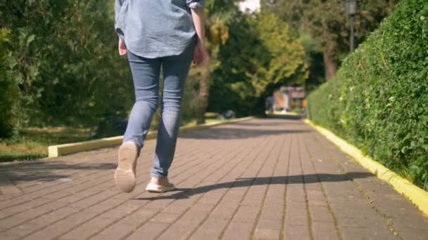 Piernas de mujer en jeans y zapatillas de deporte están en un camino pavimentado — Vídeo de stock