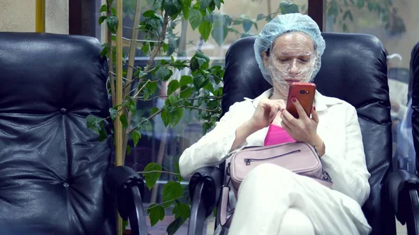 Een vrouw leest iets in een mobiele telefoon, zittend op een stoel in de gang van een cosmetologie kliniek met een film voor anesthesie op haar gezicht. — Stockfoto