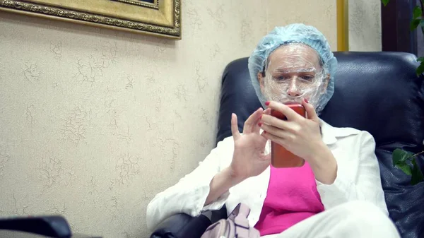 Een vrouw leest iets in een mobiele telefoon, zittend op een stoel in de gang van een cosmetologie kliniek met een film voor anesthesie op haar gezicht. — Stockfoto