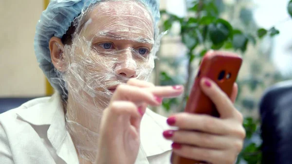 Kobieta czyta coś w telefonie komórkowym, siedząc na krześle w korytarzu kliniki kosmetologii z filmem do znieczulenia na twarzy. — Zdjęcie stockowe
