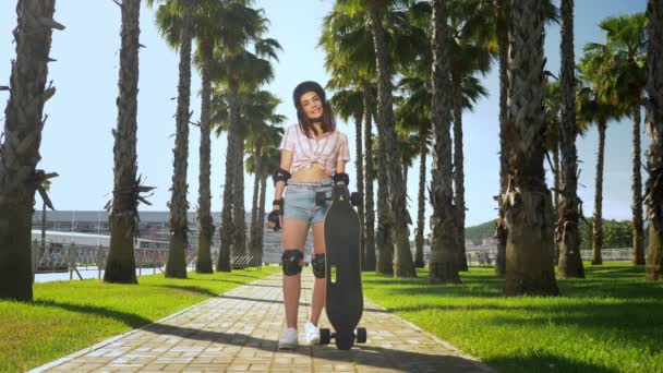 一个美丽的年轻黑发女孩，戴着头盔和溜冰鞋保护站在公园的高棕榈树的背景，手里拿着溜冰鞋，看着相机，面带微笑 — 图库视频影像