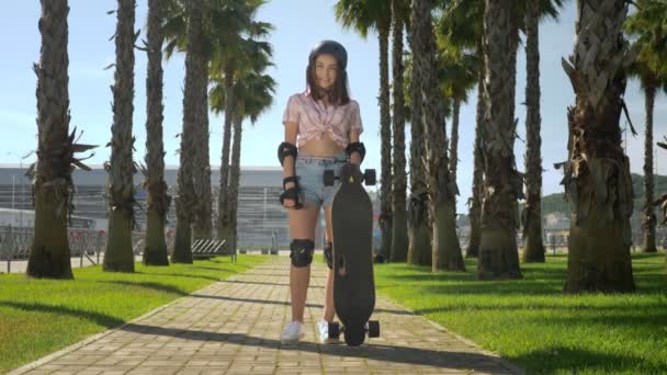 ヘルメットとスケート保護の美しい若いブルネットの女の子は、彼女の手にスケートを保持し、カメラを見て、笑顔で、高いヤシの木の背景に公園に立っています — ストック動画