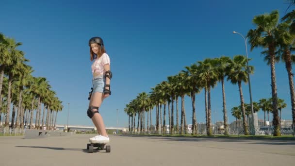 Menina montando um skate elétrico em um belo parque com palmeiras altas — Vídeo de Stock