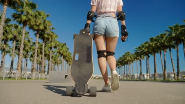 Close-up, uma menina caminha por um parque com palmeiras altas e rola um skate atrás dela. a câmera a segue — Vídeo de Stock