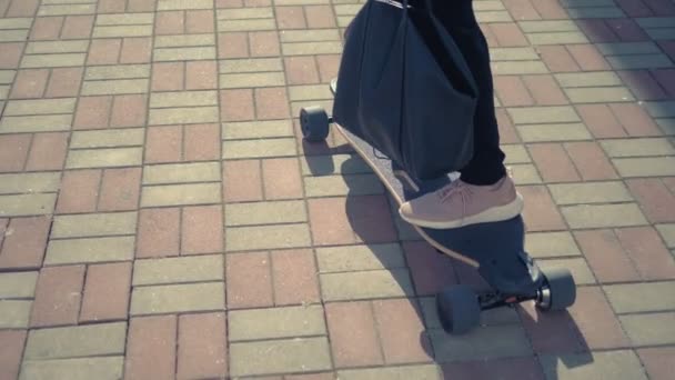 Una mujer de negocios moderna en un mono negro en zapatillas de deporte de color rosa, gafas de sol y una bolsa negra, montando un tablero eléctrico a lo largo de una calle de la ciudad. El concepto de aventuras extrañas de la gente — Vídeo de stock