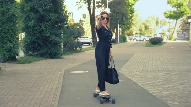 Mulher de negócios moderna em um macacão preto em tênis rosa, óculos de sol e um saco preto montado em uma placa elétrica ao longo de uma rua da cidade, representando um super homem, esticando um braço para a frente. O conceito — Vídeo de Stock