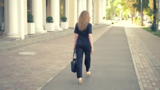 A young woman with long dark blond hair, dressed in a black jumpsuit, beige leather shoes, with a black bag walking down the street, the camera follows her, view from the back — Stock Video