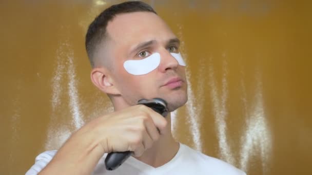 Портрет красивого мужчины в маске для глаз, который бреется электрической бритвой в ванной. Метросексуальное понятие — стоковое видео