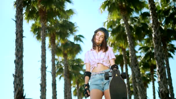 Una bella ragazza bruna con un casco e una protezione per pattini è in piedi nel parco sullo sfondo di alte palme, con un pattino in mano, guardando la fotocamera e sorridendo — Video Stock