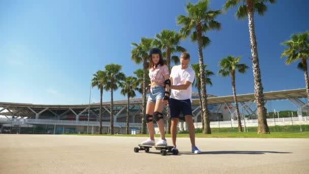 Ragazzo insegna ragazza a cavalcare uno skateboard elettrico in un parco con alte palme — Video Stock