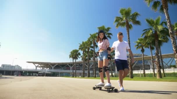 Guy lär flickan att rida en elektrisk skateboard i en park med höga palmer — Stockvideo