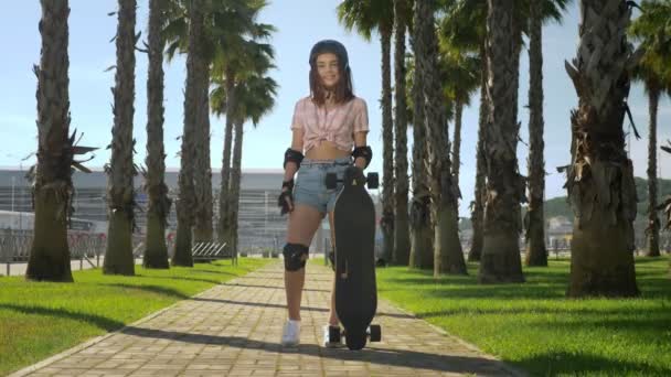 Uma linda garota morena em um capacete e proteção de patins está de pé no parque no fundo de palmeiras altas, segurando um skate nas mãos, olhando para a câmera e sorrindo — Vídeo de Stock