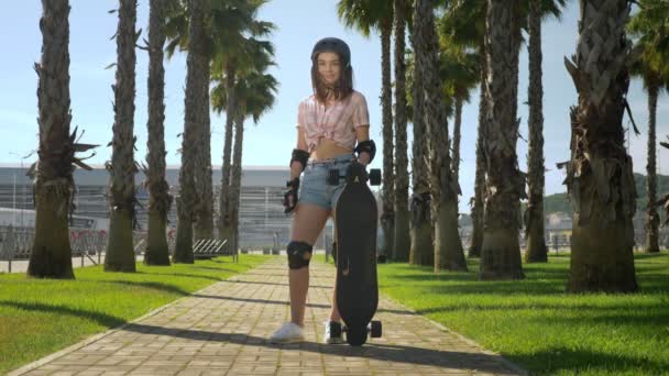 Piękna Młoda Dziewczyna Brunetka w kask i skate ochrony stoi w parku na tle wysokich Palm, trzymając skate w dłoniach, patrząc na aparat i uśmiechnięty — Wideo stockowe