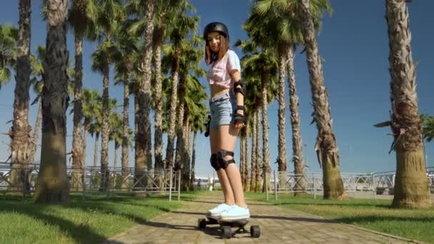 Menina montando um skate elétrico em um belo parque com palmeiras altas — Vídeo de Stock