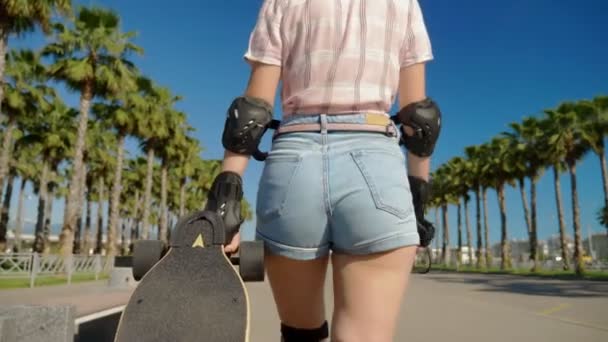 Gros plan, une fille marche à travers un parc avec de grands palmiers et roule une planche à roulettes derrière elle. la caméra la suit — Video