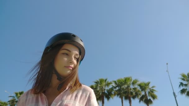 Cara de cerca, una chica con casco. chica montando un monopatín eléctrico en un hermoso parque con palmeras altas — Vídeo de stock
