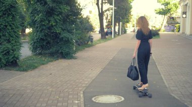 A modern business woman in a black jumpsuit in beige pumps shoes, and with a black bag riding on an electric board along a city street. Concept of strange people adventures clipart