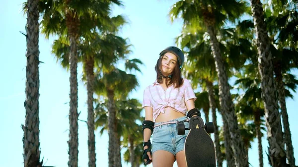 Egy gyönyörű fiatal barna lány a sisak és Skate védelem áll a parkban a háttérben a magas pálmafák, gazdaság egy korcsolyázik a kezében, nézte a kamerát, és mosolyogva — Stock Fotó