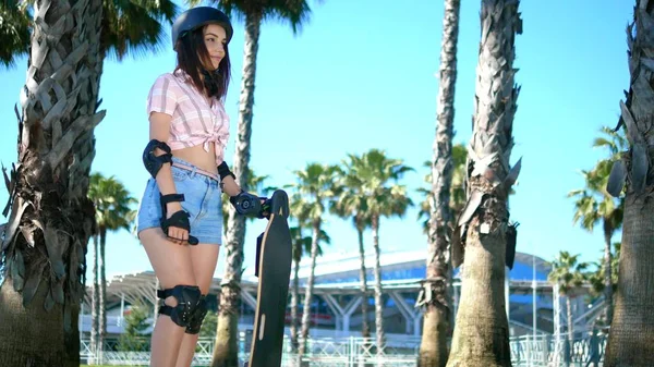 Une belle jeune fille brune avec un casque et une protection contre le patin se tient dans le parc sur le fond de hauts palmiers, tenant un patin dans ses mains — Photo