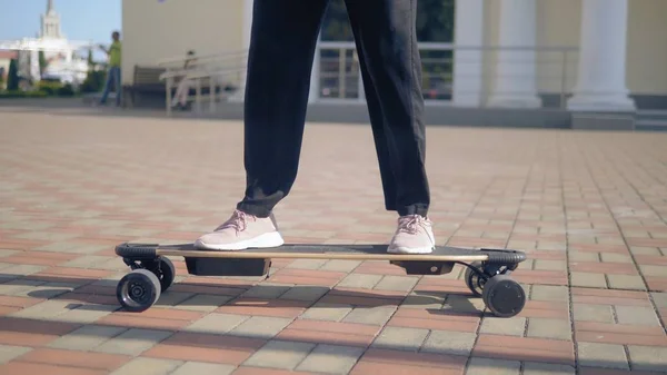 a modern business woman in a black jumpsuit in pink sneakers, sunglasses and a black bag, riding an electricboard along a city street. The concept of strange adventures of people