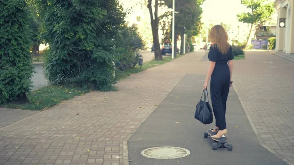 Uma mulher de negócios moderna em um macacão preto em sapatos de bombas bege, e com um saco preto montando em uma placa elétrica ao longo de uma rua da cidade. Conceito de pessoas estranhas aventuras — Fotografia de Stock