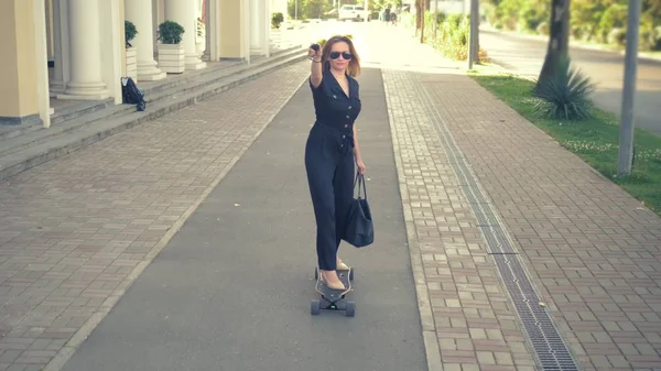 Mulher de negócios moderna em um macacão preto em sapatos bege, óculos de sol e um saco preto montado em uma placa elétrica ao longo de uma rua da cidade, representando um super homem, esticando um braço para a frente. O conceito de — Fotografia de Stock