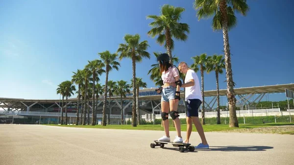 Chlap učí dívku jezdit na elektrickém skateboard v parku s vysokými palmami — Stock fotografie