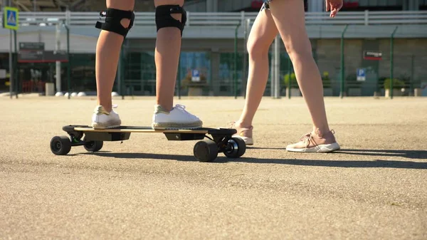 Mädchen auf einem Elektro-Skateboard. Nahaufnahme, weibliche Beine. ein paar Mädchen, die im Park spazieren gehen, ein Mädchen auf einem Elektro-Skateboard, das andere läuft nebenher und hält ihre Hand — Stockfoto