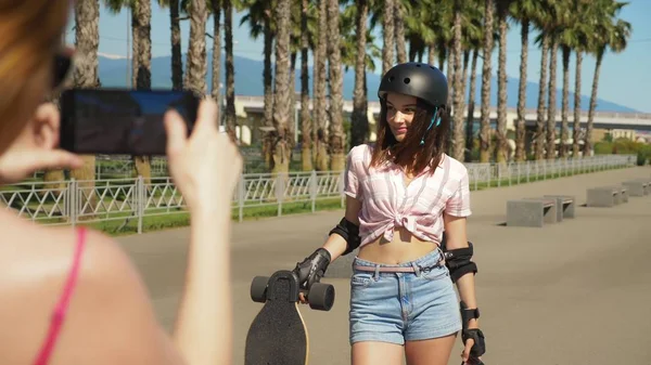 Sepasang gadis berjalan di taman, seorang gadis dengan skateboard listrik di tangannya berpose, yang lain melakukan fotonya di smartphone, konsep blog — Stok Foto