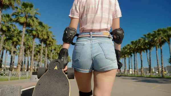 Zblízka, dívka prochází parkem s vysokými palmami a za ní vyjíždí skateboard. kamera sleduje její — Stock fotografie