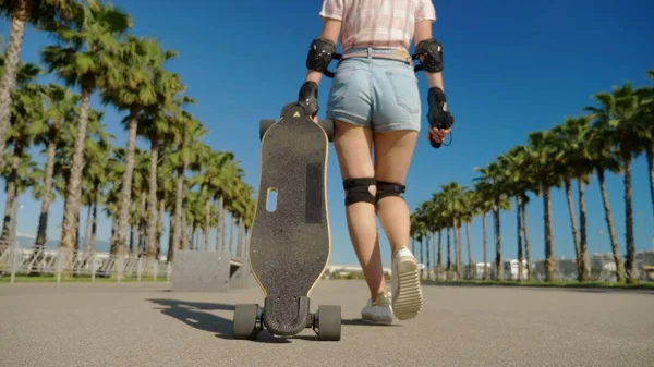 Zblízka, dívka prochází parkem s vysokými palmami a za ní vyjíždí skateboard. kamera sleduje její — Stock fotografie
