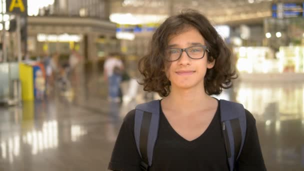 Un adolescente felizmente mira a la cámara mientras está en el aeropuerto. Fondo desenfocado — Vídeos de Stock