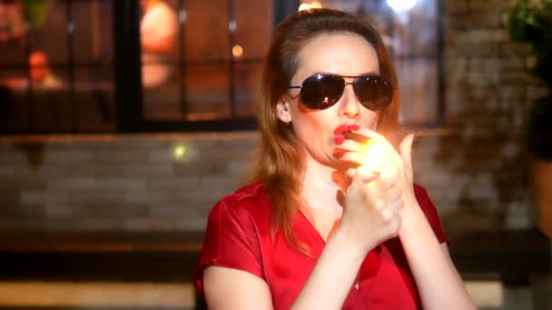 Hermosa mujer en vestido rojo con lápiz labial rojo en los labios y gafas oscuras con un cigarro sobre un fondo oscuro. produce humo espeso de la boca — Vídeos de Stock